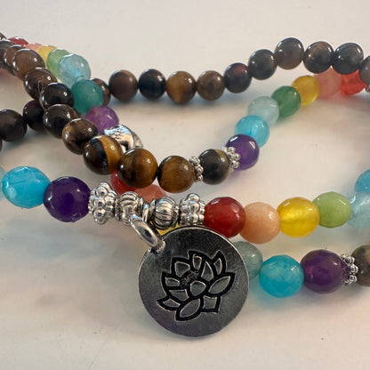 A close-up of a beaded necklace with multicolored gemstone-like round beads, silver-toned spacer beads, and a round dark gray metal pendant engraved with a lotus flower. The necklace is photographed on a plain light background.